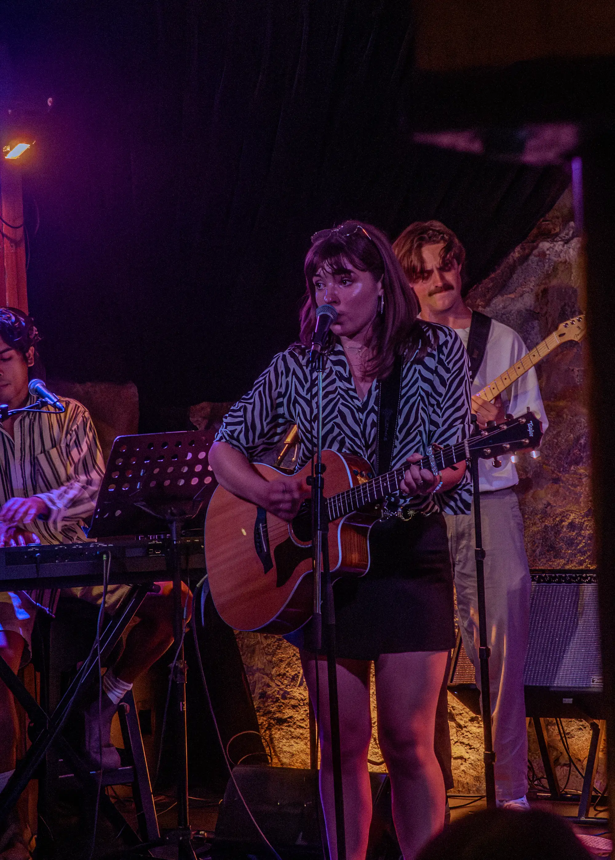 a full stage in amber light, a singer with an acoustic guitar at the microphone, keys and electric guitar behind