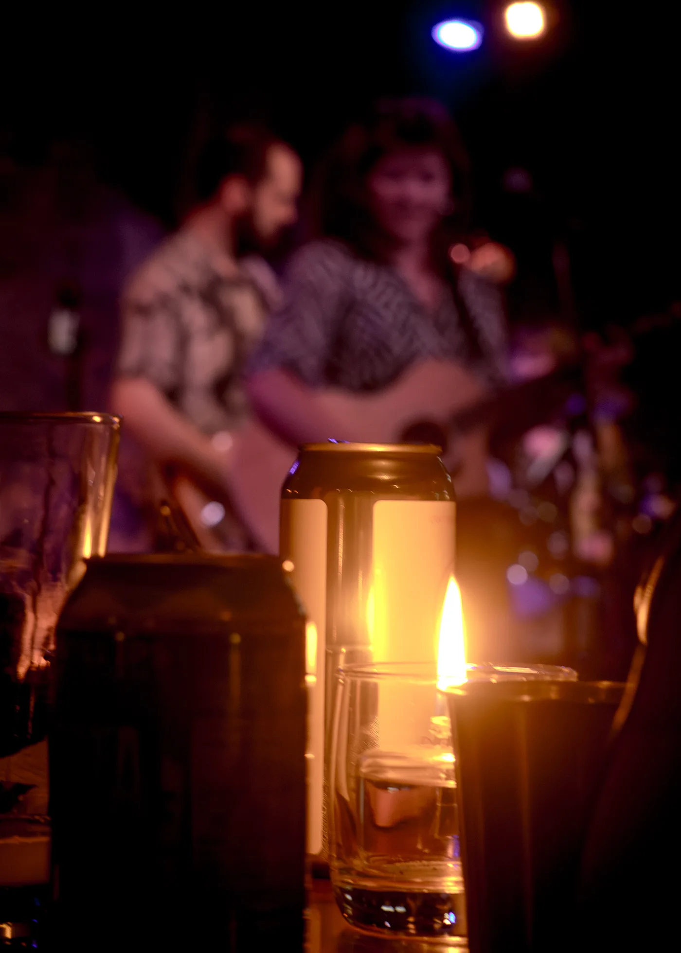 a lit candle and drinks on a table in the foreground, a duo with an acoustic guitar softly out of focus on stage behind