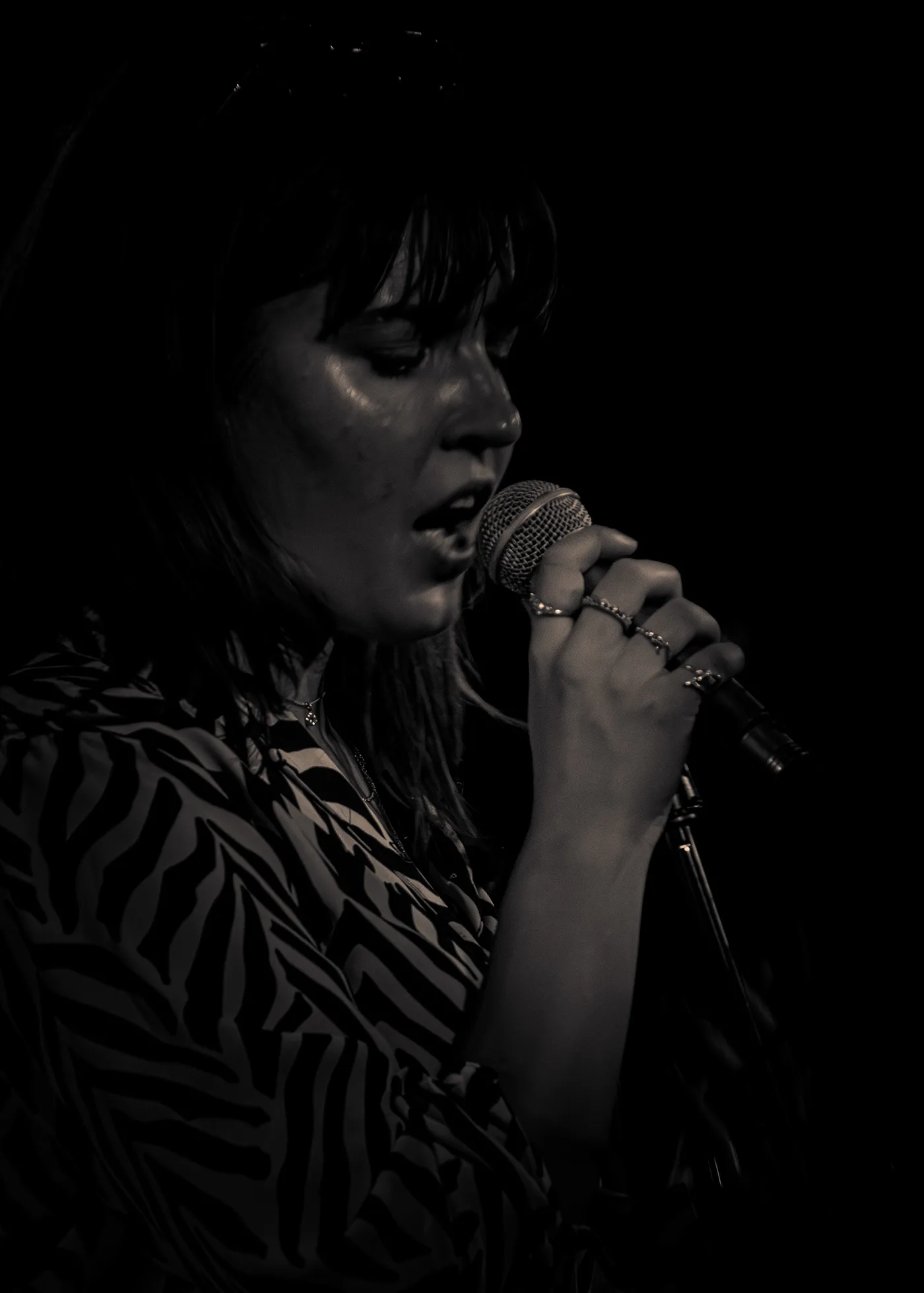a close sepia portrait of a singer at a handheld microphone, eyes lowered, rings on her fingers