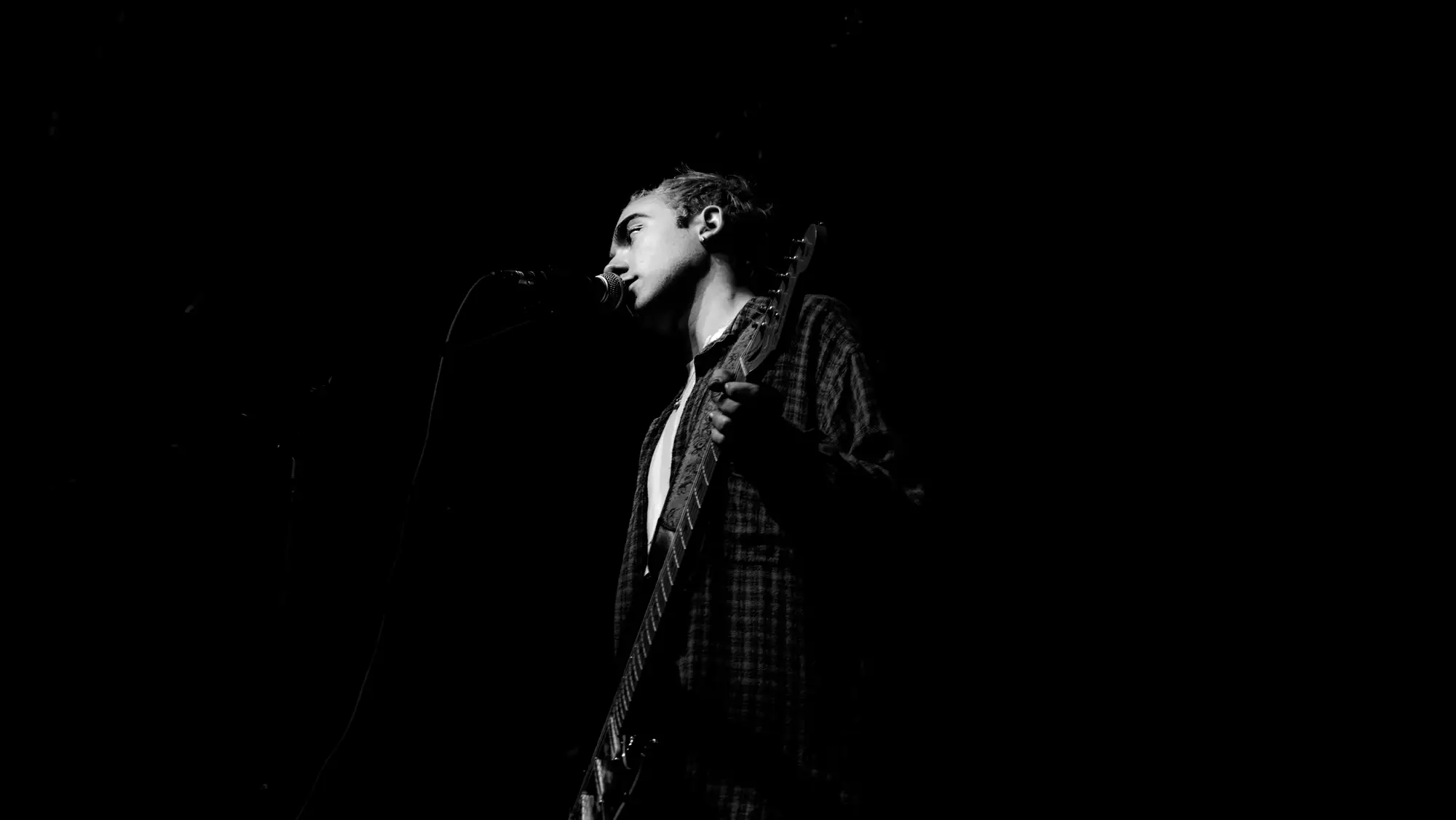 a singer in profile at the microphone, guitar slung, one shaft of light against the dark
