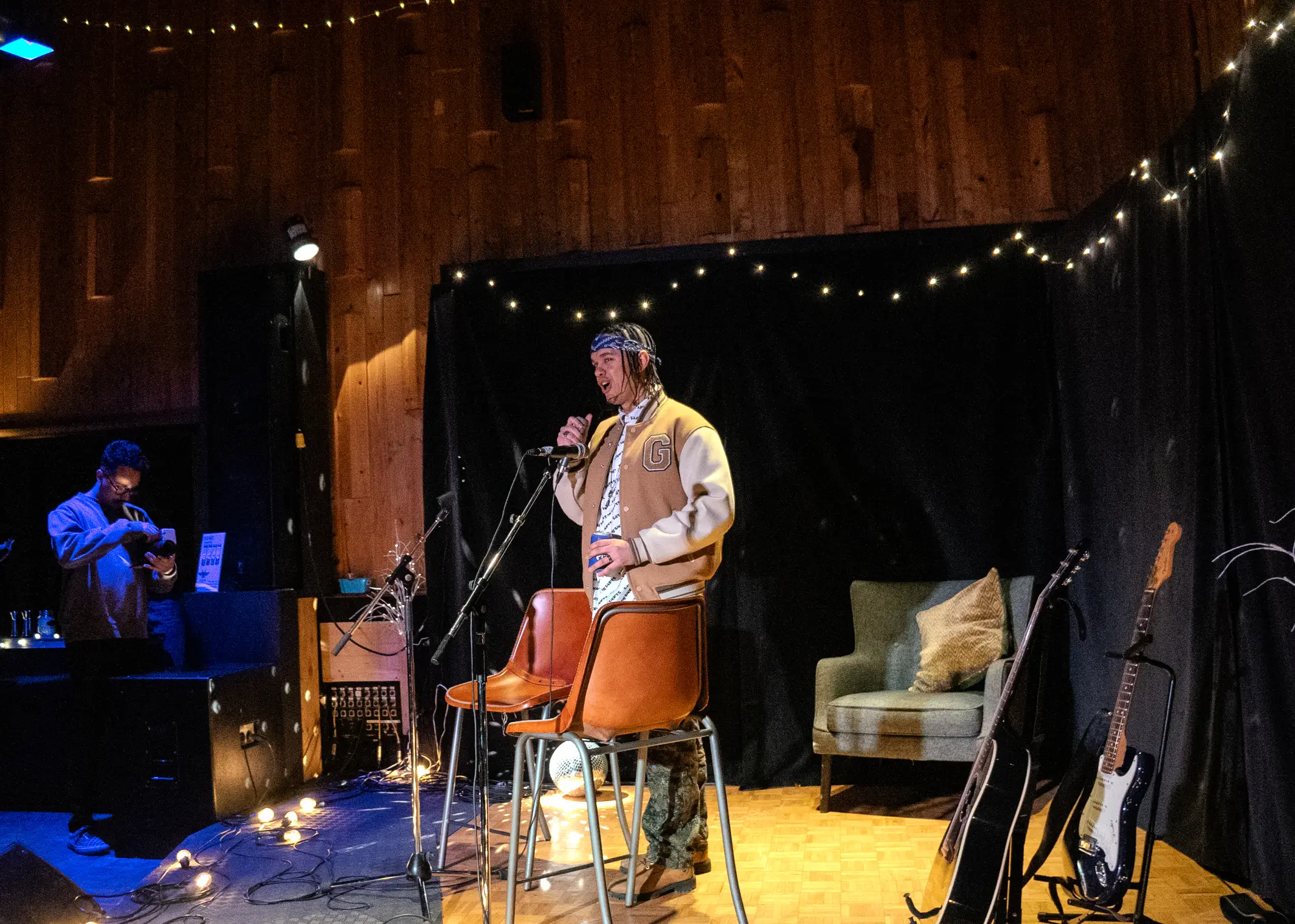 a wood-panelled hall strung with fairy lights, a singer at the microphone beside stacked chairs and a green armchair