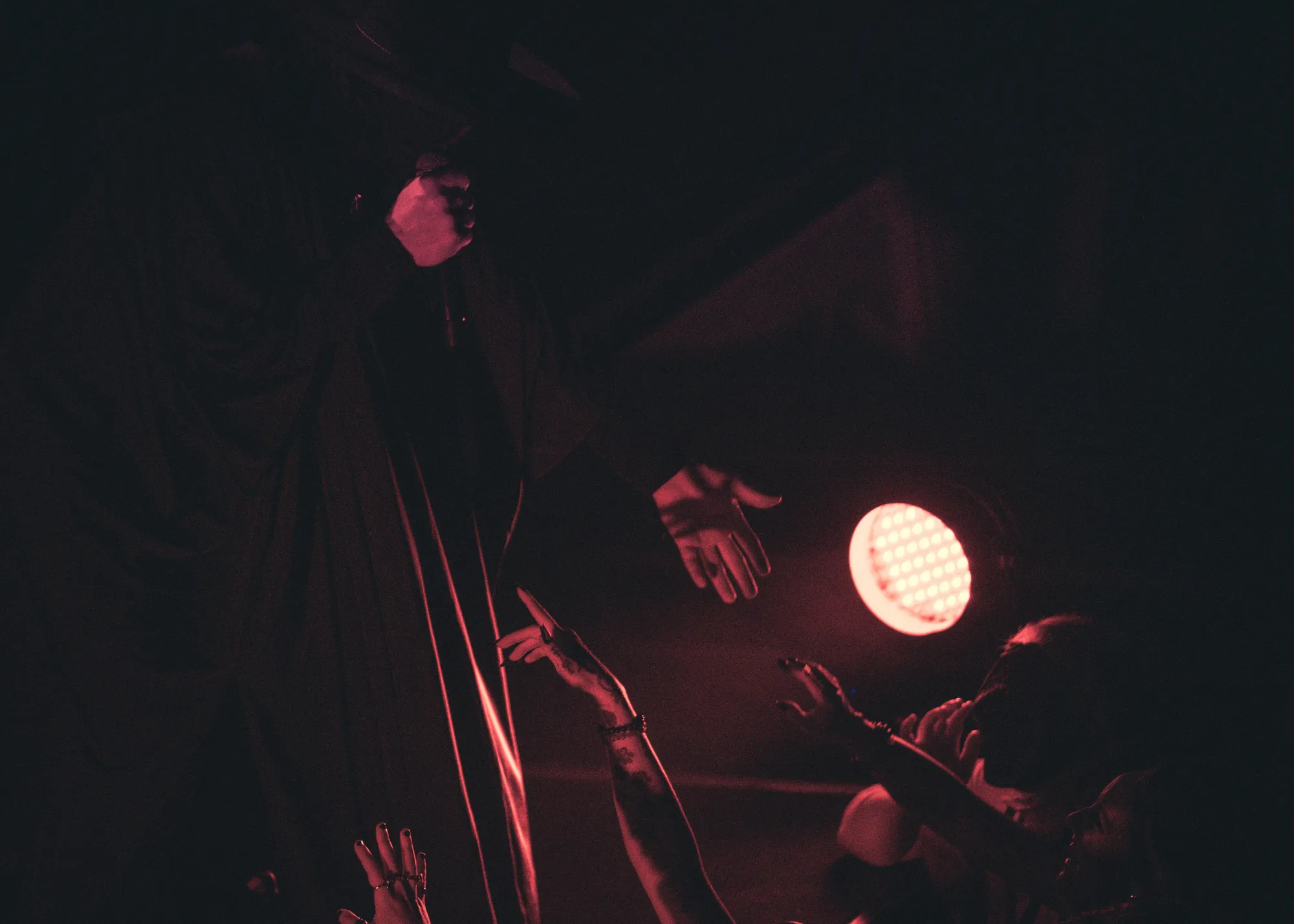 A robed figure in deep red light extends a hand over a reaching crowd