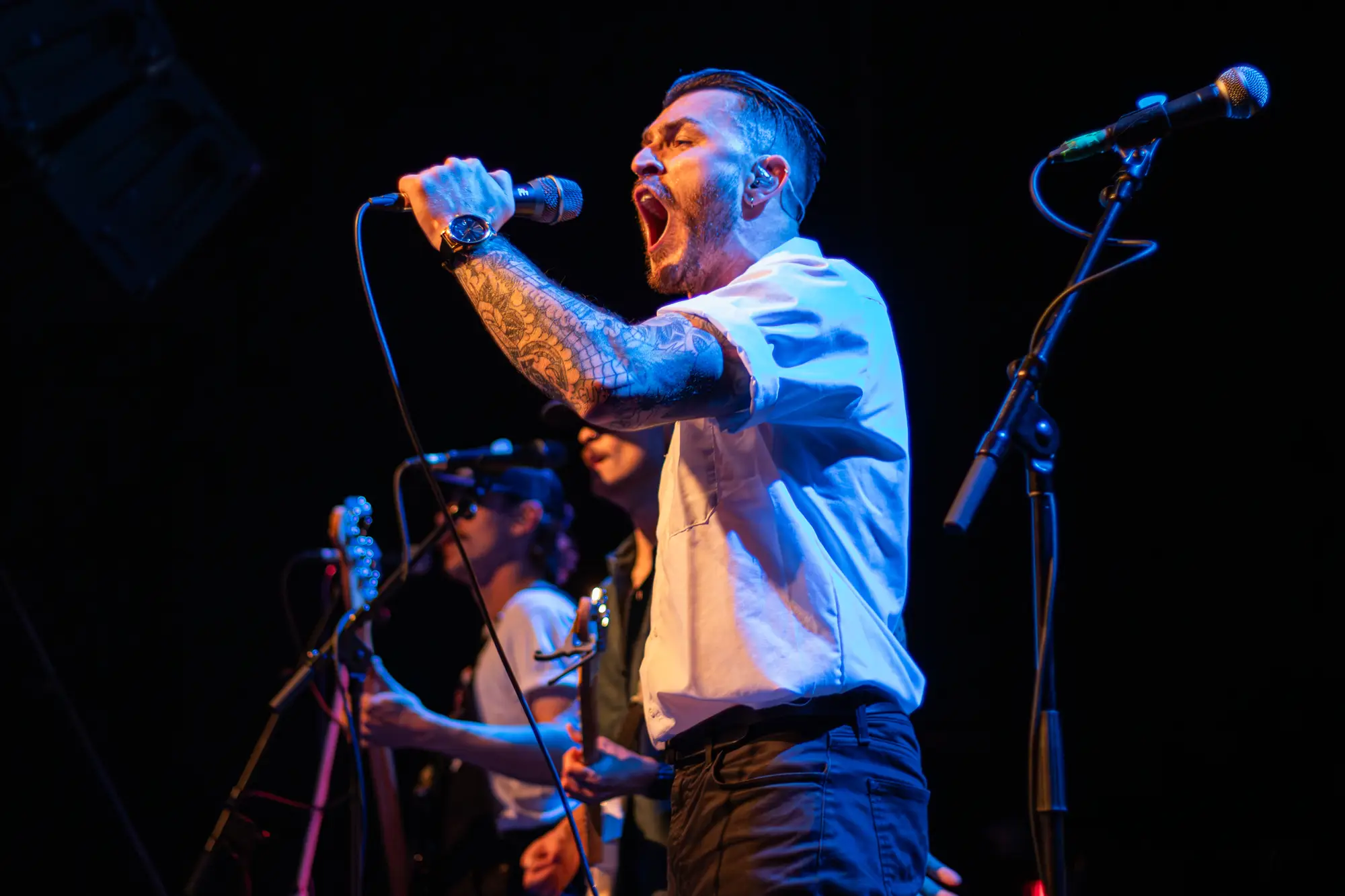 A vocalist in a white shirt screaming into a hand-held mic under blue light