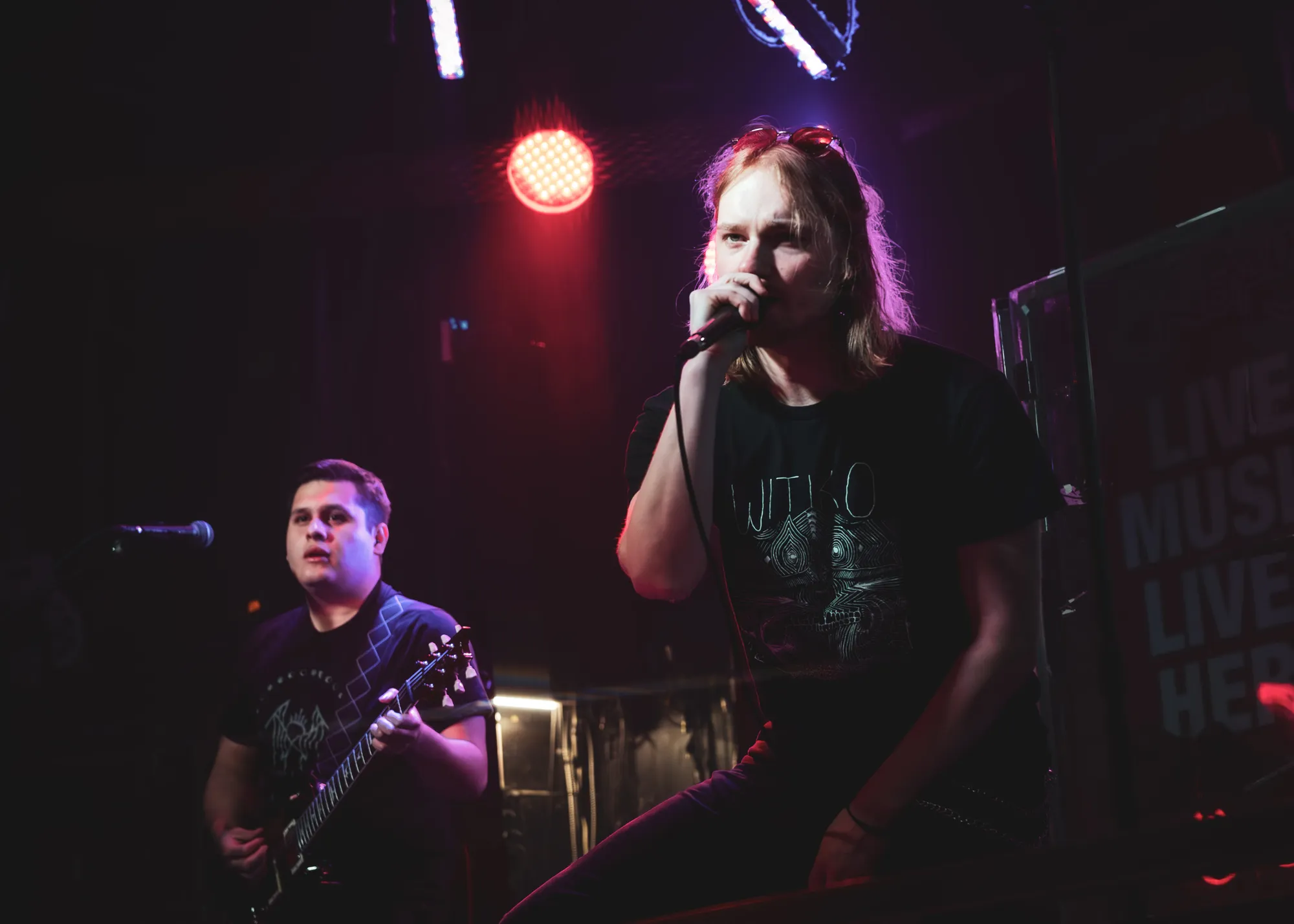 Vocalist gripping the mic under a red spotlight while the guitarist watches