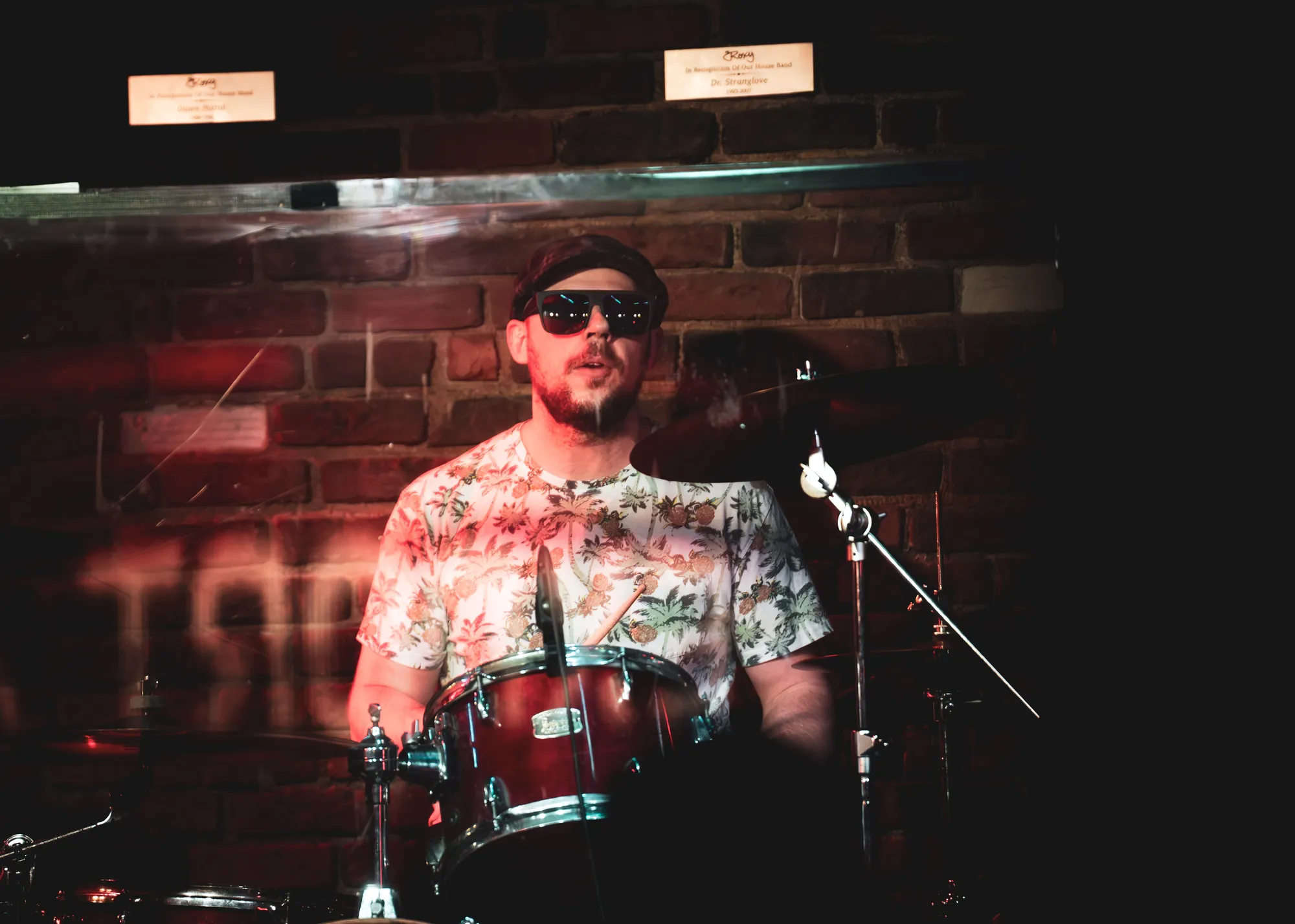 Drummer in sunglasses and a floral shirt in front of the Roxy's brick wall