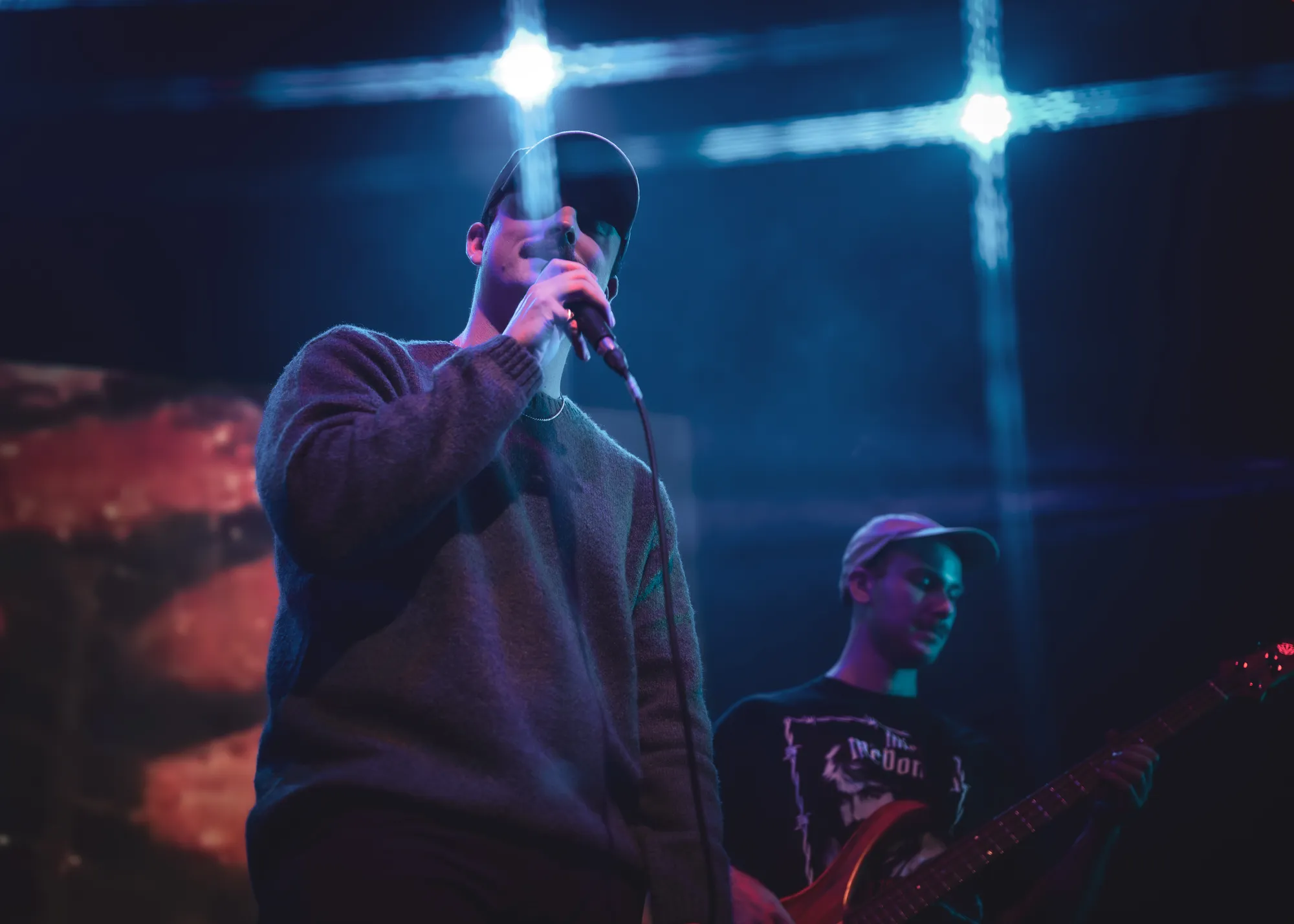 Singer in a cap under cross-shaped blue lights, guitarist behind